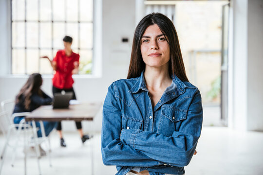 Portrait of a young female businesswoman with arms folded in her new office, behind her two colleagues working together - Successful millennial of a self-confident start-up - Concept of teamwork