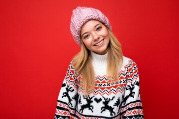 Photo of beautiful happy smiling young blonde woman isolated over red background wall wearing winter sweater and trendy pink hat looking at camera