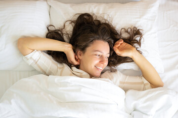 Pretty brunette young woman waking up lying in white comfortable bed, stretching arms raising hands up after sleep well, wearing pajamas. Smiling looking in distance, planning new day. Good morning.