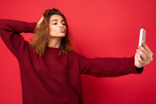 Attractive young blogger woman with curly hair wearing dark red sweater isolated on red background wall holding and using smart phone looking at telephone screen and taking selfie and giving kiss