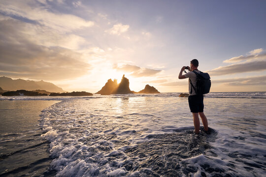 Man during photographing landscape with cliff. Young photographer on beach at beautiful sunset. Tenerife, Canary Islands, Spain.