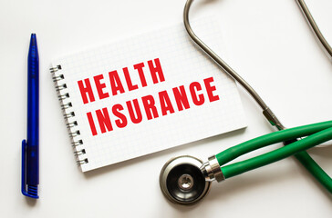 HEALTH INSURANCE is written in a notebook on a white table next to pen and a stethoscope.