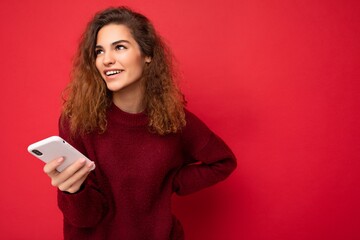 Photo of pretty young woman with curly hair wearing dark red sweater isolated on red background holding mobile phone looking to the side and laughing