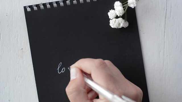 The hand writes "love you" in white ink on black paper . Top view.