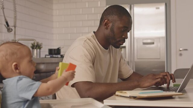 Locked-down Of Occupied African Man Working At Kitchen Table, Typing On Laptop Computer, His Mixed-Race Baby Sitting In High Chair Next To Him Playing, Woman Walking And Checking On Family Members