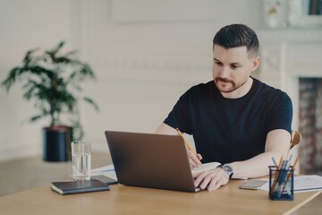 Serious bearded man writes informative notes while watching webinar on laptop computer works remotely dressed in casual t shirt poses at workplace against home office interior. Modern workspace