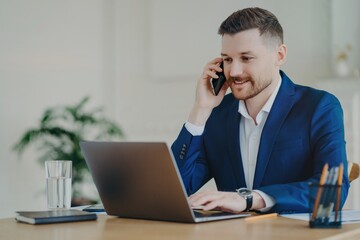 Smiling young attractive businessman talking on phone in front of laptop