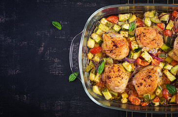 Baked chicken thighs, zucchini and vegetables in a baking dish on a dark table. Top view, overhead