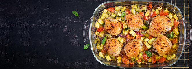Baked chicken thighs, zucchini and vegetables in a baking dish on a dark table. Top view, overhead, banner