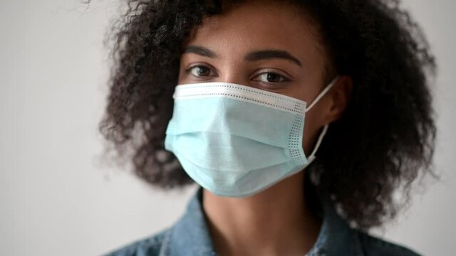 Close-up of african-american curly brunette girl, stands indoors with a protective medical mask on her face and looks directly into the camera. Quarantine precautions, healthcare concept