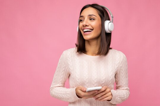 Photo of beautiful joyful smiling young woman wearing stylish casual clothes isolated over background wall holding and using mobile phone wearing white bluetooth headphones listening to music and
