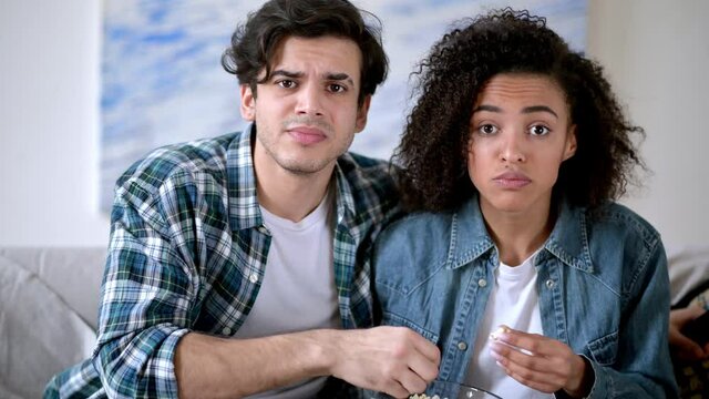 Young mixed race family couple sitting at home on sofa in casual clothes, watching tv show, exciting movie or football match, eating popcorn, worried, spending weekend together at home,emotional faces