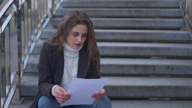Young brunette Caucasian sad woman reading letter tearing paper sitting on urban city stairs outdoors. Portrait of stressed depressed angry millennial on spring autumn day. Slow motion