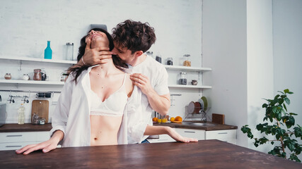man hugging sensual woman in white shirt and bra near table in kitchen