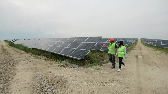 Diverse Engineers In Special Uniform Walking Through Solar Farm. The Rows Of Solar Batteries. Green Energy Concept. Solar Panel Field.