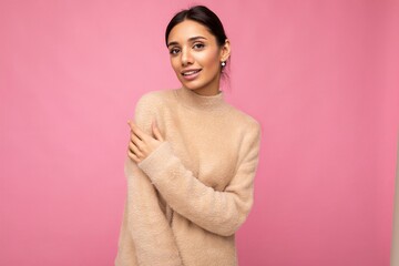 Portrait of beautiful positive cheerful cute smiling young brunette woman in casual beige sweater isolated on pink background with copy space and looking at camera