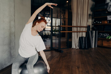 Sportive red haired woman sitting on big fitball and stretching body before workout in gym