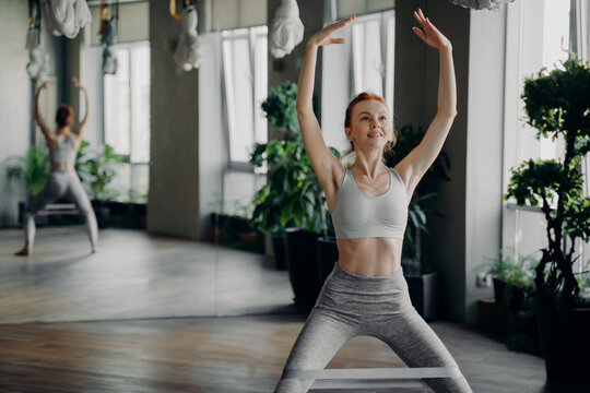 Athletic slender redhead fitness instructor sits doing squats with pilates rubber resistance band on legs