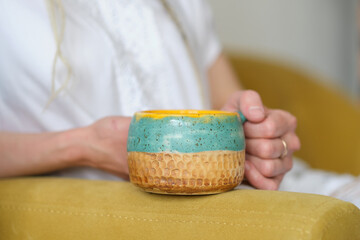 unrecognizable woman in white clothes sits on a yellow sofa and holds a yellow handmade cup.