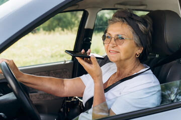 Portrait of joyful mature woman with glasses talking on speakerphone on mobile phone while driving a car. Trendy smiling senior woman holding wheel and using smartphone. Active lifestyle pensioner