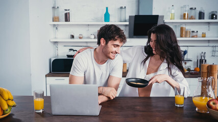 sexy woman in unbuttoned shirt showing frying pan to man near laptop in kitchen