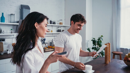 pretty woman holding smartphone near smiling man reading morning newspaper