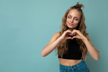 Photo of smiling positive adorable young cute nice blonde curly lady with sincere emotions wearing black top isolated over blue background with empty space and showing heart gesture. Love concept