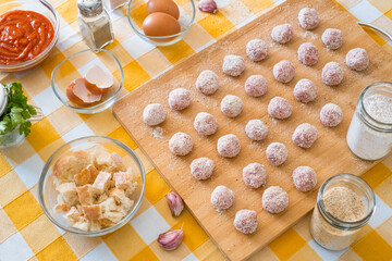 Above view of kitchen table with meatballs ready for pan, wooden cutting board and raw ingredients