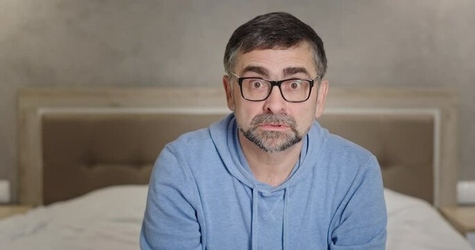 Mad mature man looking at camera.Angry middle aged male gesticulating and looking at camera in rage while sitting on bed in bedroom at home