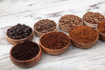 Overhead view of five varieties of roasted coffee beans with their freshly ground counterparts in small bowls on a wooden wooden background.