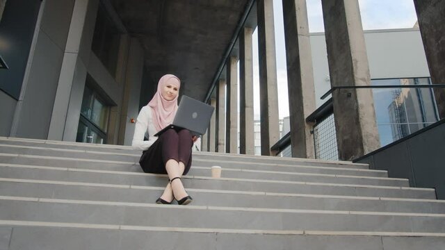 Portrait of attractive arabian woman in elegant clothes and hijab sitting on stairs with modern laptop on knees. Charming woman in stylish elegant clothes and hijab. Muslim female working on distance