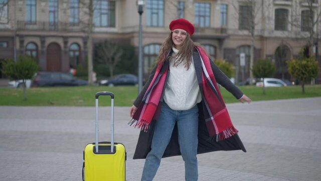 Wide shot of happy excited stylish young woman dancing spinning in slow motion on urban street with yellow travel bag on sidewalk. Cheerful Caucasian tourist enjoying vacations trip outdoors