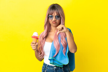 Young mixed race woman with pink hair holding ice cream isolated on yellow background showing thumb down with negative expression