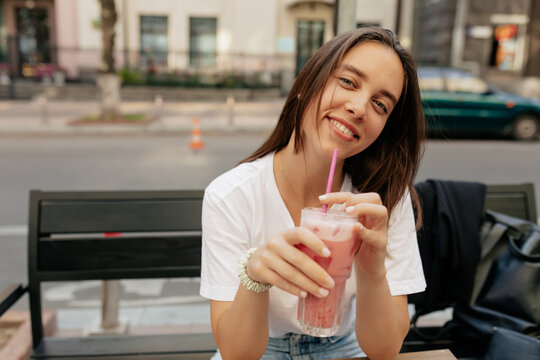 Charming young lady with lovely smile, white t-shirt and jeans sitting outdoors on city cafe terrace, drinking lemonade and smiling