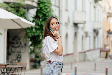 Fancy girl with brown hair wearing white t-shirt is talking on the phone while walking in the city center