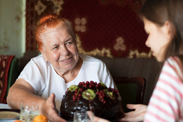 An elderly woman sits at the table and with a smile looks at a young girl,a granddaughter,who wishes her grandmother a happy birthday with a large chocolate cake. Focus on woman. Girl and cake blurred