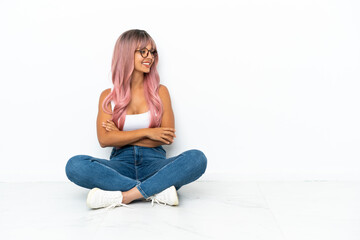 Young mixed race woman with pink hair sitting on the floor isolated on white background with arms crossed and happy