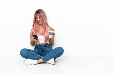 Young mixed race woman with pink hair sitting on the floor isolated on white background holding coffee to take away and a mobile