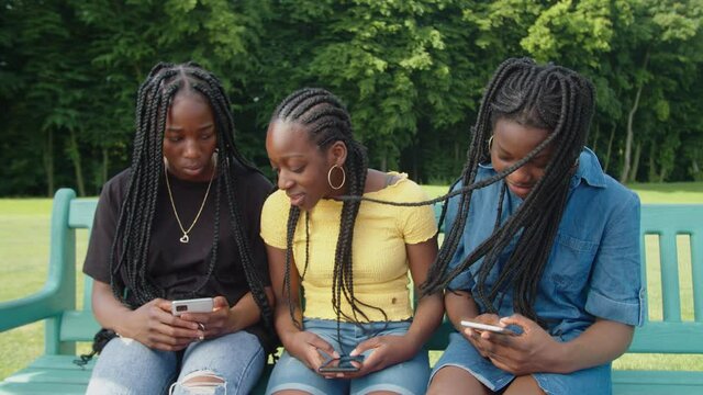 Cheerful positive lovely black teenage girls with braided hair sitting on park bench, networking and browsing online social media content on smartphones while relaxing together in summer nature.