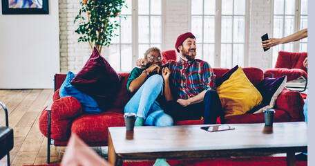 Cropped woman showing modern smartphone to cheerful couple in love rejoicing at comfortable sofa in apartment, joyful marriage laughing on couch while reading funny text publication on mobile phone