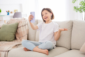 Photo of sweet charming school girl wear white t-shirt smiling sitting sofa talking modern device indoors house home room
