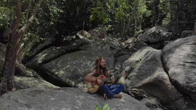 Man with dreadlocks sitted on a rock playing guitare - drone shot