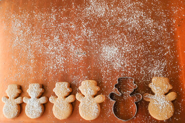 Minimalist Christmas background with copy space: a row of homemade snowman cookies and a cookie cutter on copper background covered with icing sugar (snow)
