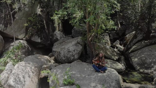 Man with dreadlocks sitted on a rock playing guitare - drone shot