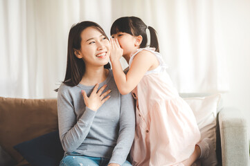 Little Asian daughter girl whispering a secret to happy smiling mother in living room