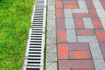 grating of drainage system rainwater in the park at the sidewalk from a stone tilea with a green lawn, landscaped a city garden engineering systems close-up, nobody.