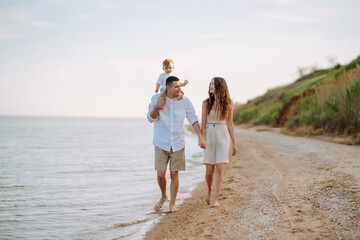 Cheerful young family with little baby boy spending time together on the beach. Father, mother and child against the background of the blue sea and sky. Family, childhood, active lifestyle concept.