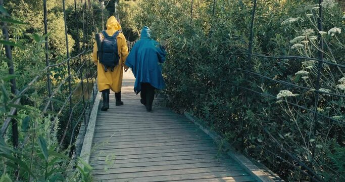 Man and woman hikers with backpacks in yellow and blue raincoats walk on wooden suspension bridge over river. Concept of travel, hiking, lifestyle and outdoor recreation. Sun is shining. From back