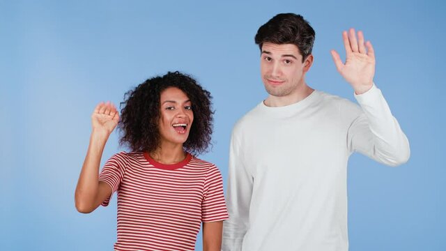 Interracial friendly couple - white man and african woman waving hands - goodbye, chao, adios. Parting, say bye to camera on blue studio background.