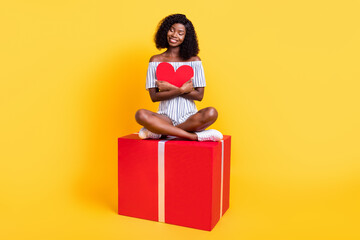 Portrait of attractive cheerful dreamy girl sitting on big box hugging heart card isolated over bright yellow color background